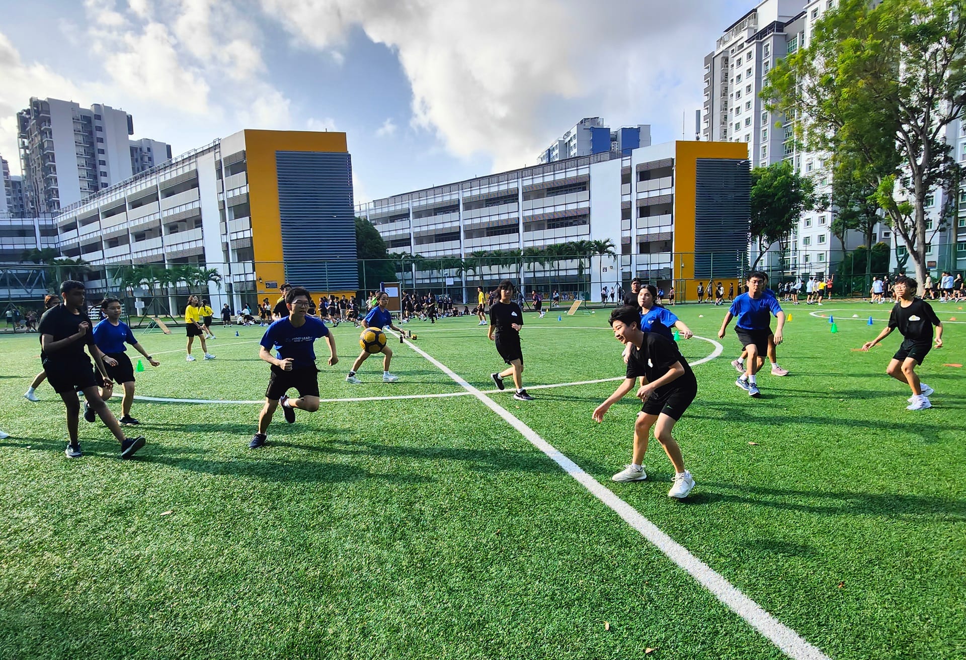 Students play a game on a green artificial turf field with school buildings in the background.